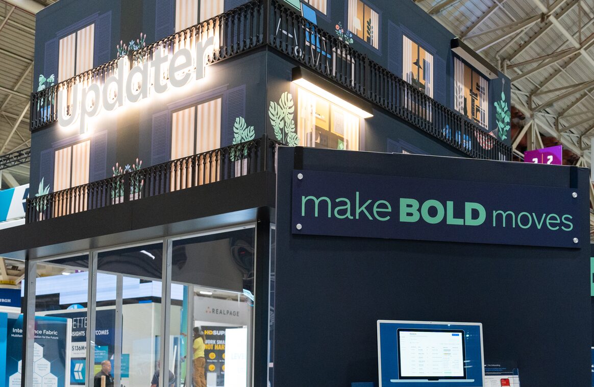 The two-storey Updater booth at a trade show, its lit logo above a 'make BOLD moves' sign