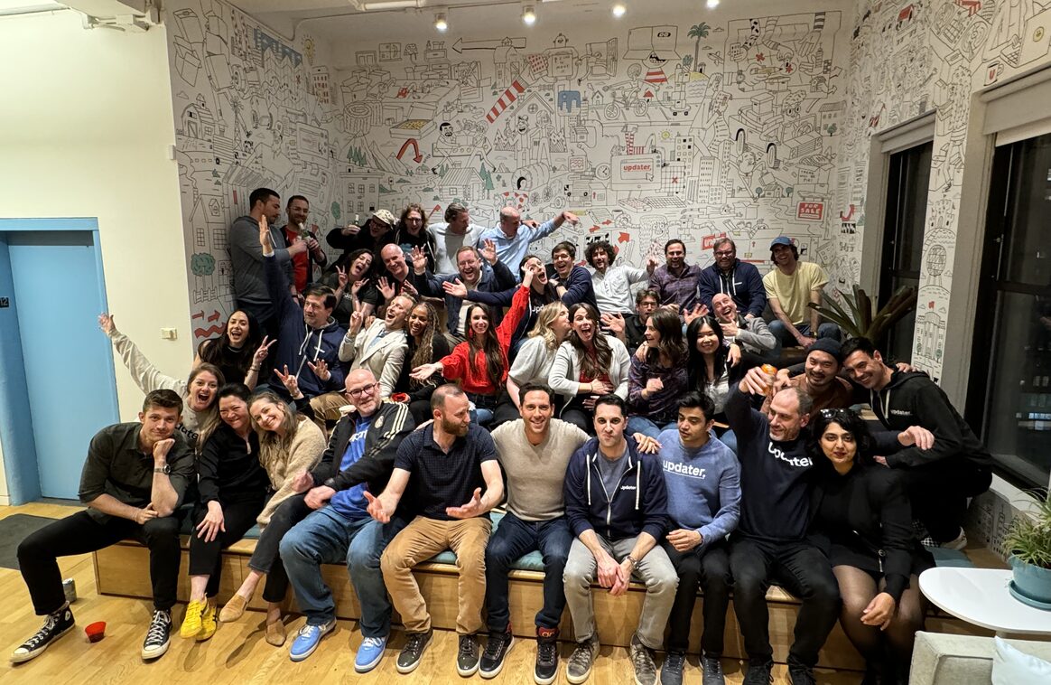 Dozens of Updater colleagues posing together in front of a hand-drawn city mural at the office