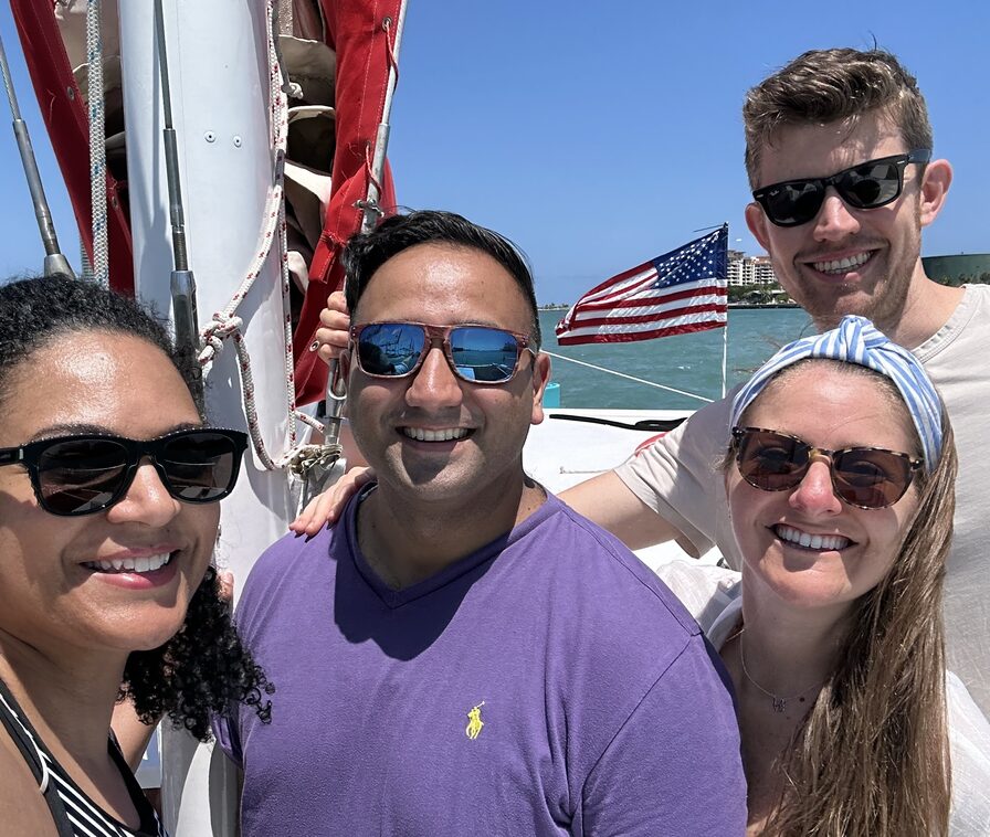Four Updater colleagues smiling together on a sailboat, an American flag and the shoreline behind them