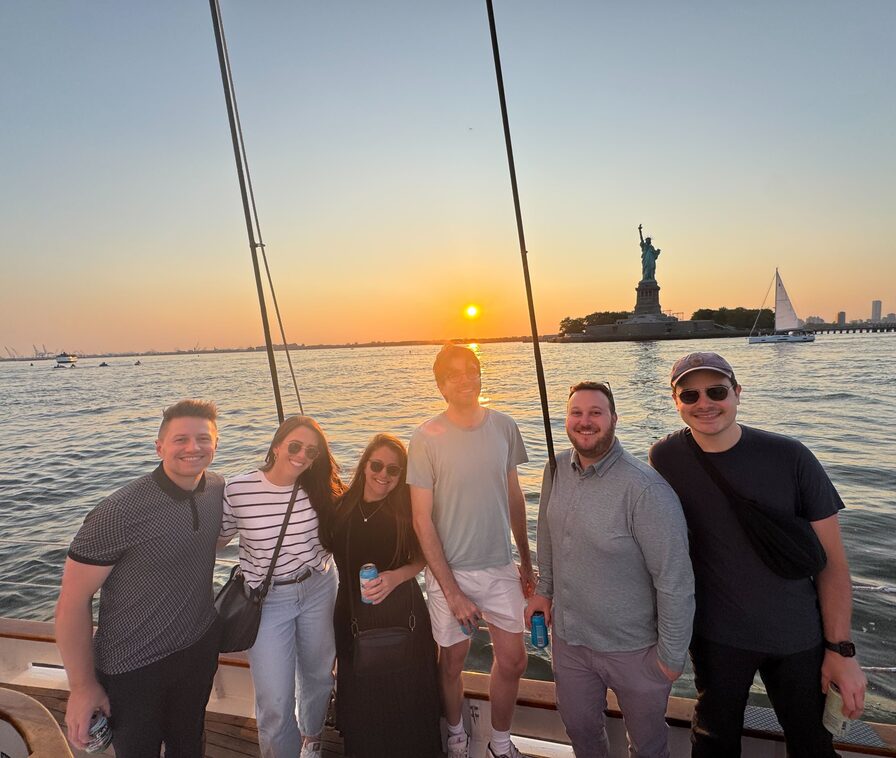 Six Updater colleagues on a sailboat deck at sunset, the Statue of Liberty and New York Harbor behind them