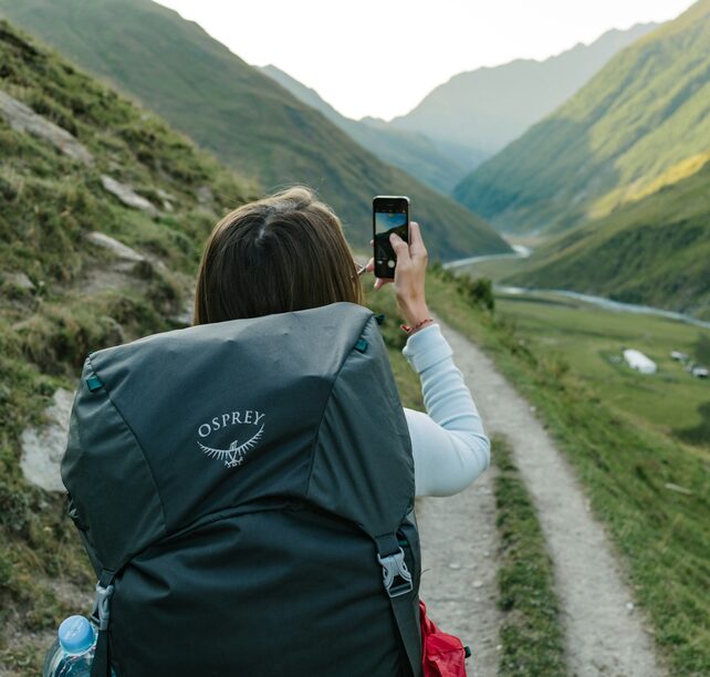 A hiker with a large backpack photographing a mountain valley from a trail